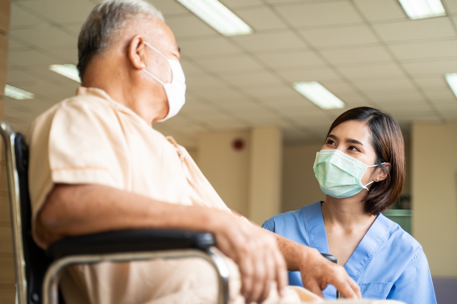 A Personal Support Worker wearing a mask and scrubs kneels beside an elderly patient in a wheelchair, who is also wearing a mask.